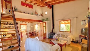 A spacious living area with a high ceiling, wooden beams, a fireplace, a sofa, a dining table, and a staircase. - Ca'n Paner - One Bedroom Villa, Sleeps 2 (Mallorca)