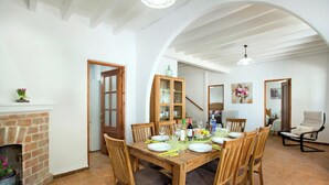 A dining area with a wooden table set for four, surrounded by chairs. A brick fireplace is visible to the left, and a wooden cabinet with glass doors is in the background. - Talati de Dalt - Five Bedroom Villa, Sleeps 8 (Alaior)