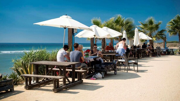 A beachside café with wooden tables and white umbrellas, overlooking the ocean.