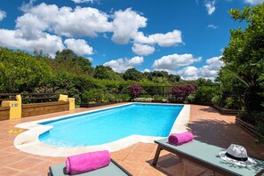 A rectangular swimming pool with a tiled deck, surrounded by lush greenery and a clear blue sky.