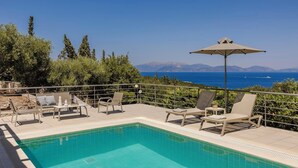 Uma área de piscina com espreguiçadeiras e um guarda-sol, com vista para um mar azul e montanhas ao longe.