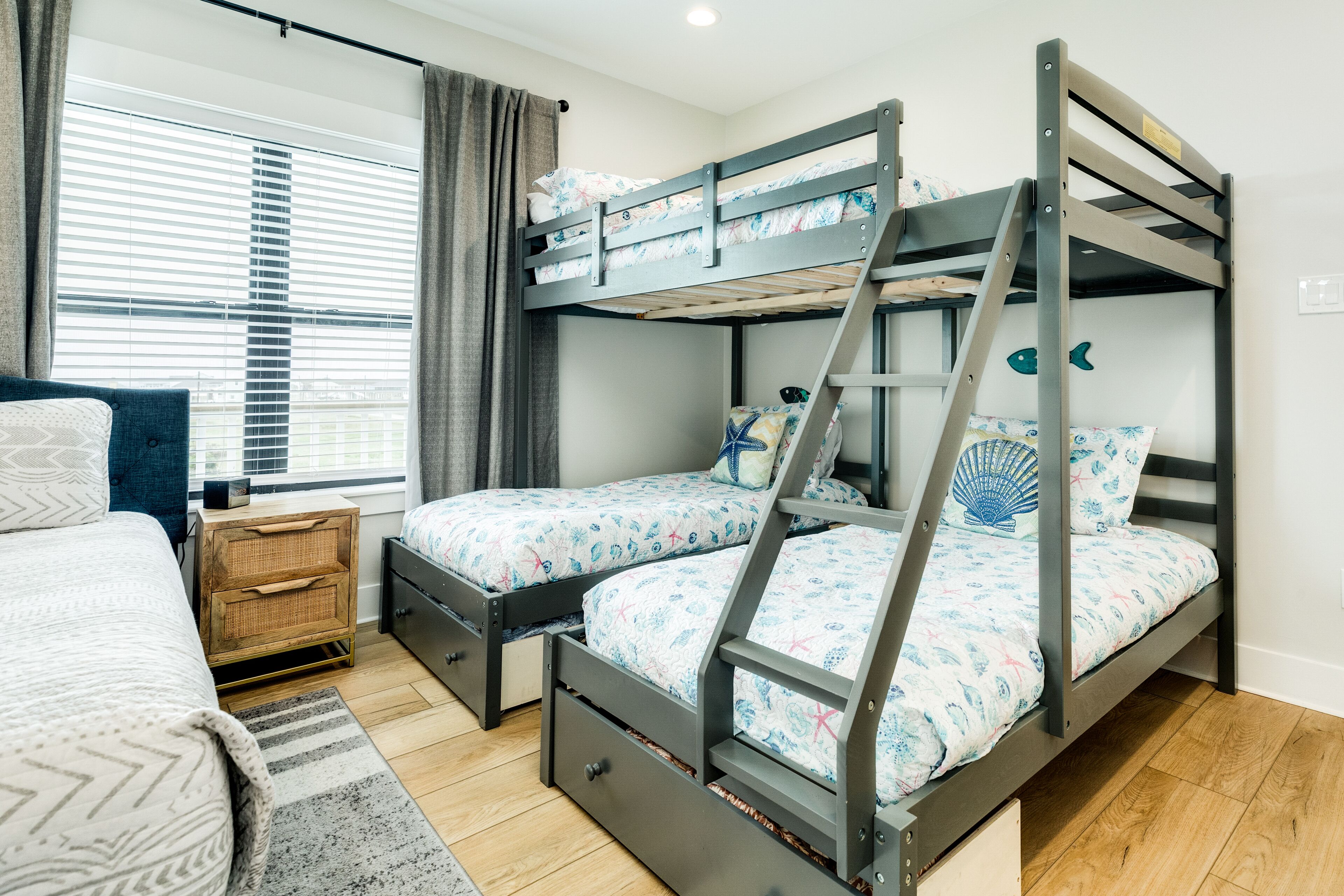 Un lit superposé avec une échelle, une table de chevet, une commode, un lit recouvert d’une couvre-lit à motifs et une fenêtre avec des stores.