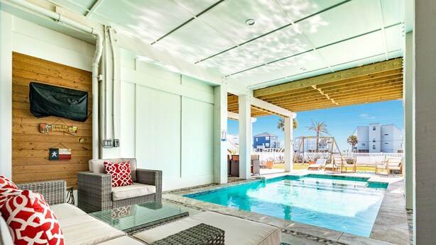 Area piscina con mobili in vimini, un tavolo in vetro e vista sulla piscina e sugli edifici circostanti.