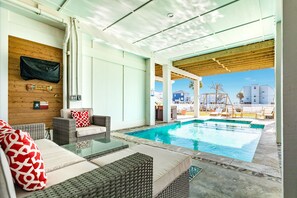 A poolside area with wicker furniture, a glass table, and a view of the pool and surrounding buildings. - Private Pool! Luxe Designer Interior, Fenced Yard, Large Bedrooms, Deck & Patio (Galveston)