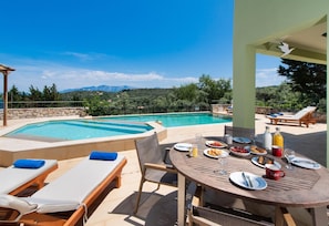 Área de refeições junto à piscina, com mesa posta para dois, com vista para uma piscina e uma paisagem montanhosa.