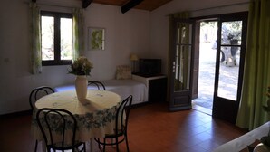 Un'area pranzo con un tavolo rotondo, sedie, un vaso con fiori, un letto con televisore e una porta che dà sull'esterno.