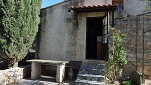 Un edificio rustico in pietra con una porta di legno, un piccolo spazio esterno con posti a sedere e un albero nelle vicinanze.