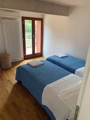 Een slaapkamer met twee bedden, een houten vloer, een raam met jaloezieën en een deur naar buiten.