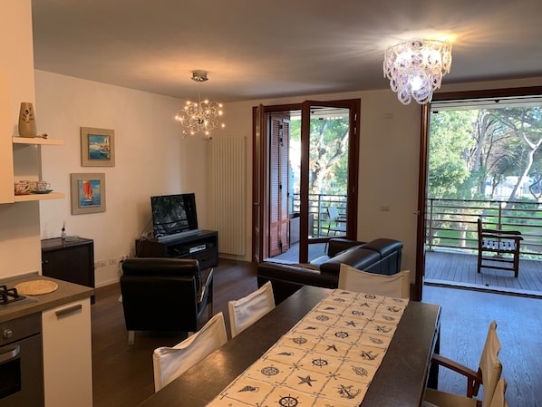 Een moderne woonkamer met een eethoek, een televisie en een balkon met uitzicht op bomen.