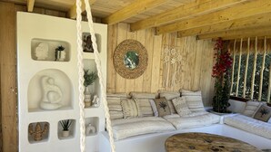 A cozy room with a wooden ceiling, a white sofa with patterned cushions, a round wooden coffee table, and a wall with decorative items including a woven basket and wall art. - Mas de Sophie (Châteaurenard)