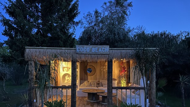 A cozy, thatched-roof structure labeled "Coco Beach" with a wooden bench and cushions, surrounded by a garden with white gravel and plants. - Mas de Sophie (Châteaurenard)