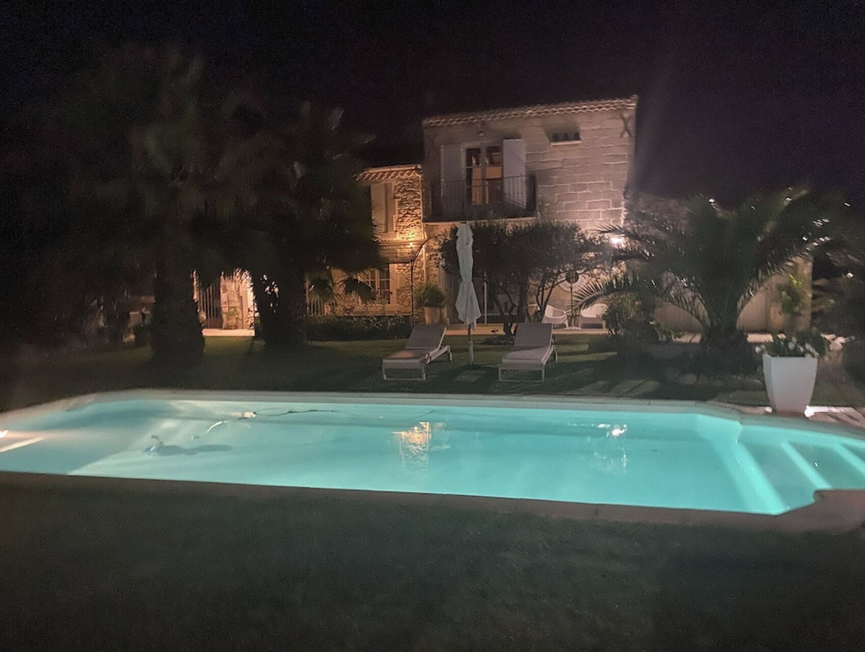 A swimming pool with lounge chairs and umbrellas, surrounded by trees and a building with stone walls and lights.