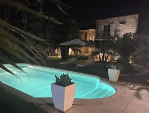 Ein Swimmingpool mit zwei Liegestühlen und einem großen Sonnenschirm, umgeben von Palmen und einem Steinhaus im Hintergrund.