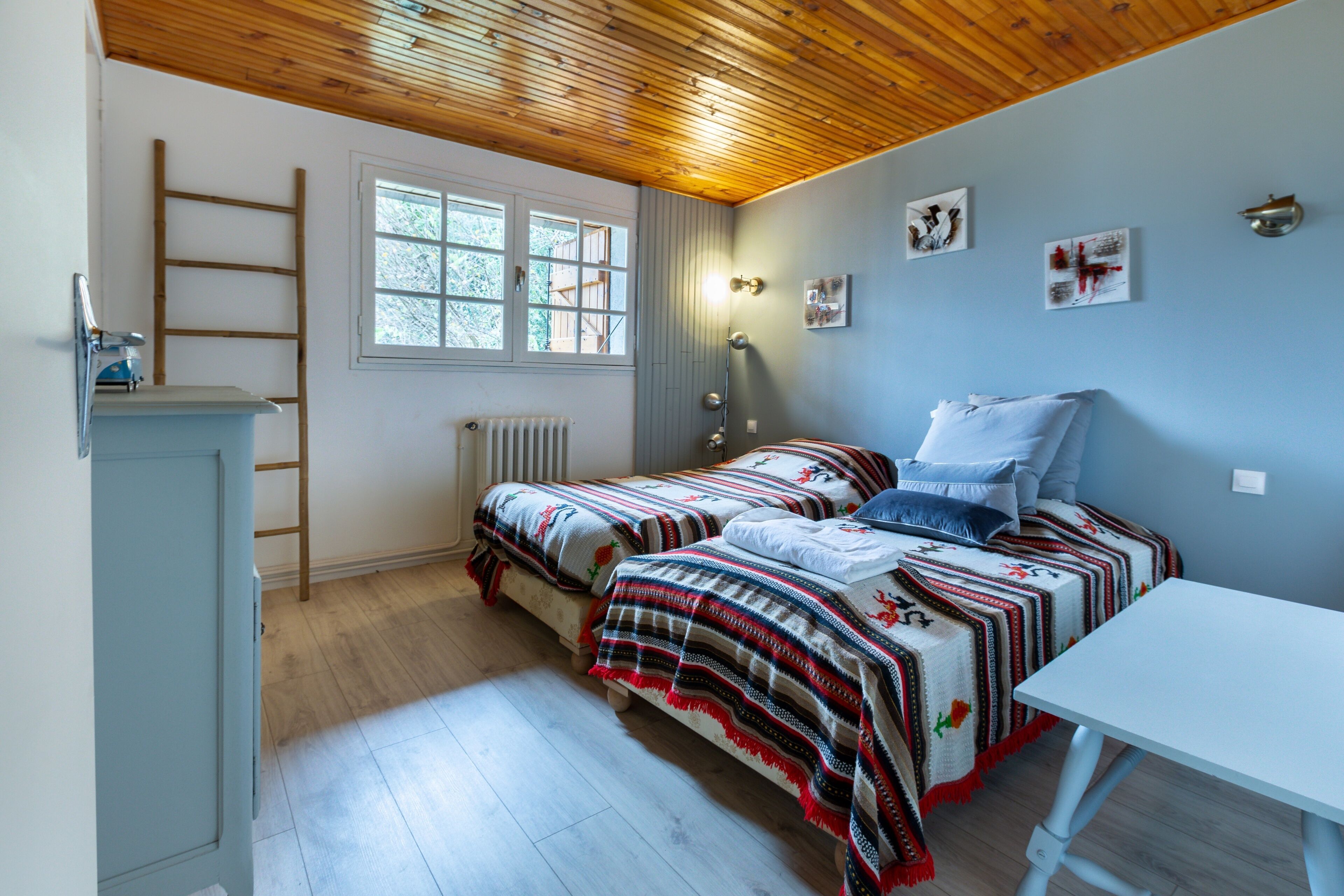 A bedroom with two beds, a wooden ceiling, a window, a radiator, and a white table.