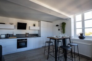 Una cocina moderna con armarios blancos, horno integrado, comedor con mesa y taburetes de madera, y ventana que proporciona luz natural.