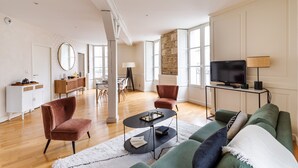 Una sala de estar moderna con un sofá verde, dos sillones, una mesa de centro y un televisor sobre un mueble bajo. Hay una zona de comedor con una mesa y sillas, además de una cocina con armarios y una estufa.