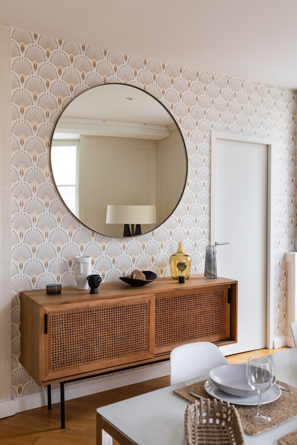 A dining area with a wooden buffet, a round mirror, and a patterned wallpaper.
