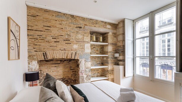 Un dormitorio con pared de piedra, una cama con almohadas, una mesita de noche, una lámpara y una ventana con vista a edificios.
