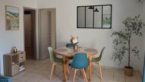 Uma sala de jantar com uma mesa redonda, quatro cadeiras, um lustre pendurado, um espelho fixado na parede e um vaso com planta.