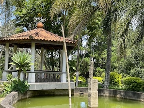 Um gazebo com telhado de azulejos, rodeado por um pequeno lago e vegetação exuberante.