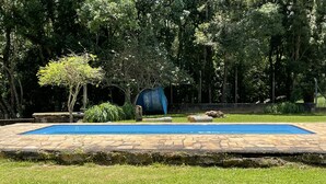 Una piscina rectangular con borde de piedra, rodeada de césped y árboles.