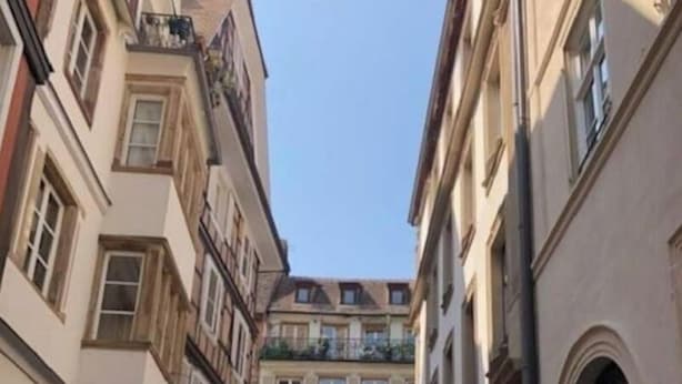 Une rue étroite, bordée de bâtiments des deux côtés, avec des terrasses de café et un ciel dégagé.