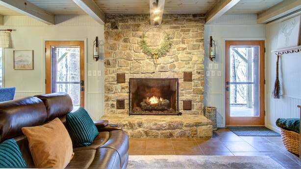A stone fireplace with a fire, a leather sofa, and a stone wall. - Beech Mtn: Walk to Slopes, Sleeps 9, Pet Friendly, Mountain Views, Cozy Retreat (Beech Mountain)