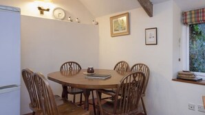 A dining area with a round wooden table and six chairs, a white refrigerator, and a window with a view of greenery.