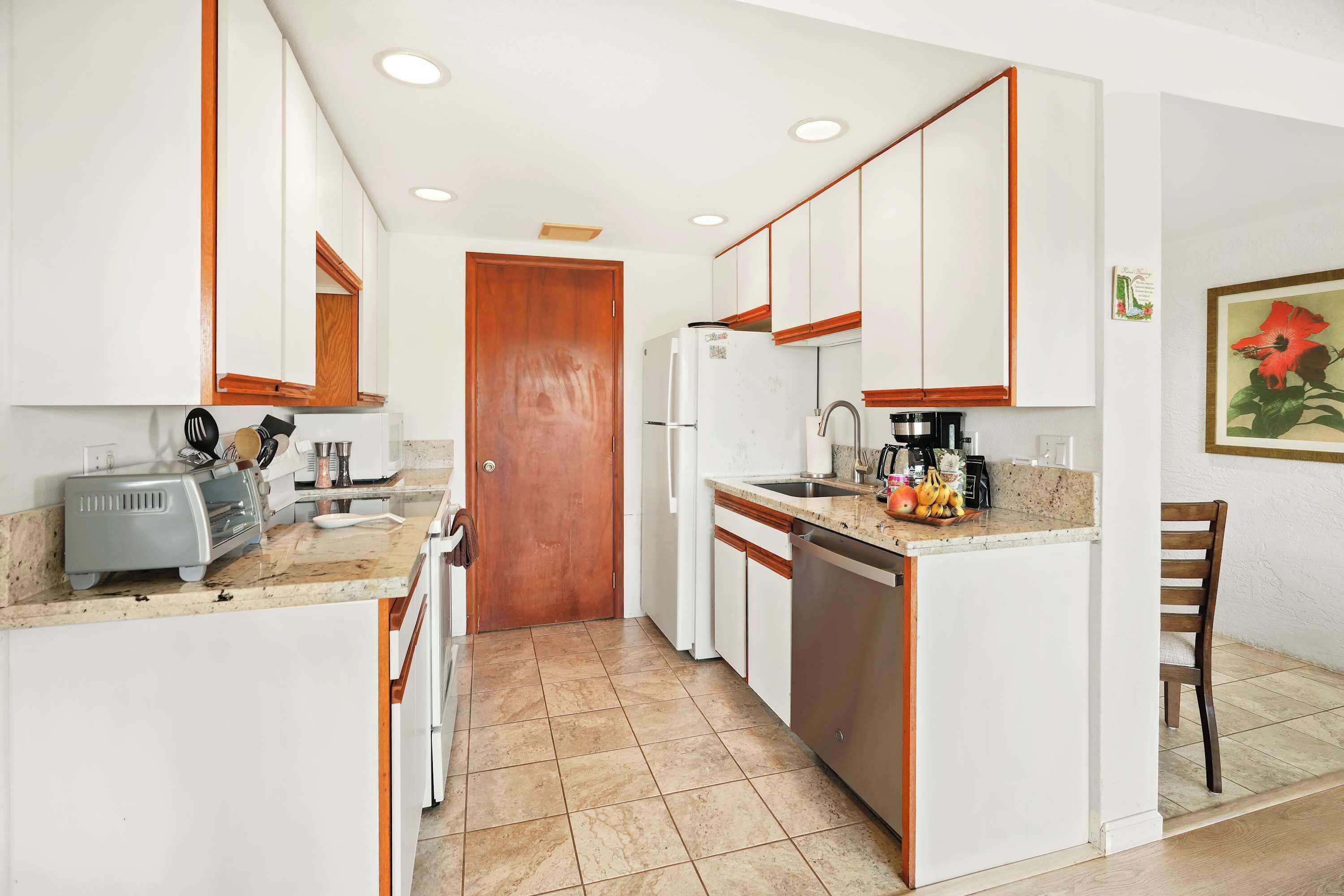 A compact kitchen with white cabinets, a wooden door, a refrigerator, a sink, and a countertop with various items.