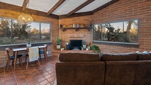 A living room with a brick fireplace, a television, and a sofa. There is a dining area with a table and chairs, and large windows offering a view of the outdoors. - Spacious casita with desert & mountain views ⛰ (Tucson)