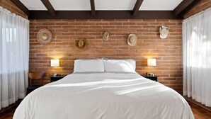 A hotel room with a large bed, two bedside tables with lamps, a brick wall, and decorative hats. - Spacious casita with desert & mountain views ⛰ (Tucson)