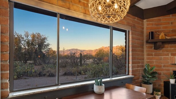 A dining area with a large window overlooking a desert landscape, a wooden table, and potted plants. - Spacious casita with desert & mountain views ⛰ (Tucson)