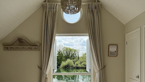 A bedroom with a large bed, a bench, a chandelier, and a view of a lake through a window. - Peaceful Lakefront Escape w/ Deck & Views (South Cerney)