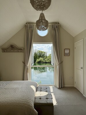 Un dormitorio con una cama grande, un banco, una araña de techo y vistas a un lago a través de una ventana.
