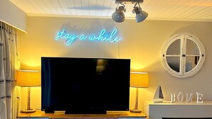A living room with a flat-screen TV, a neon sign reading "stay a while", a fireplace, and a round mirror on the wall. - Peaceful Lakefront Escape w/ Deck & Views (South Cerney)