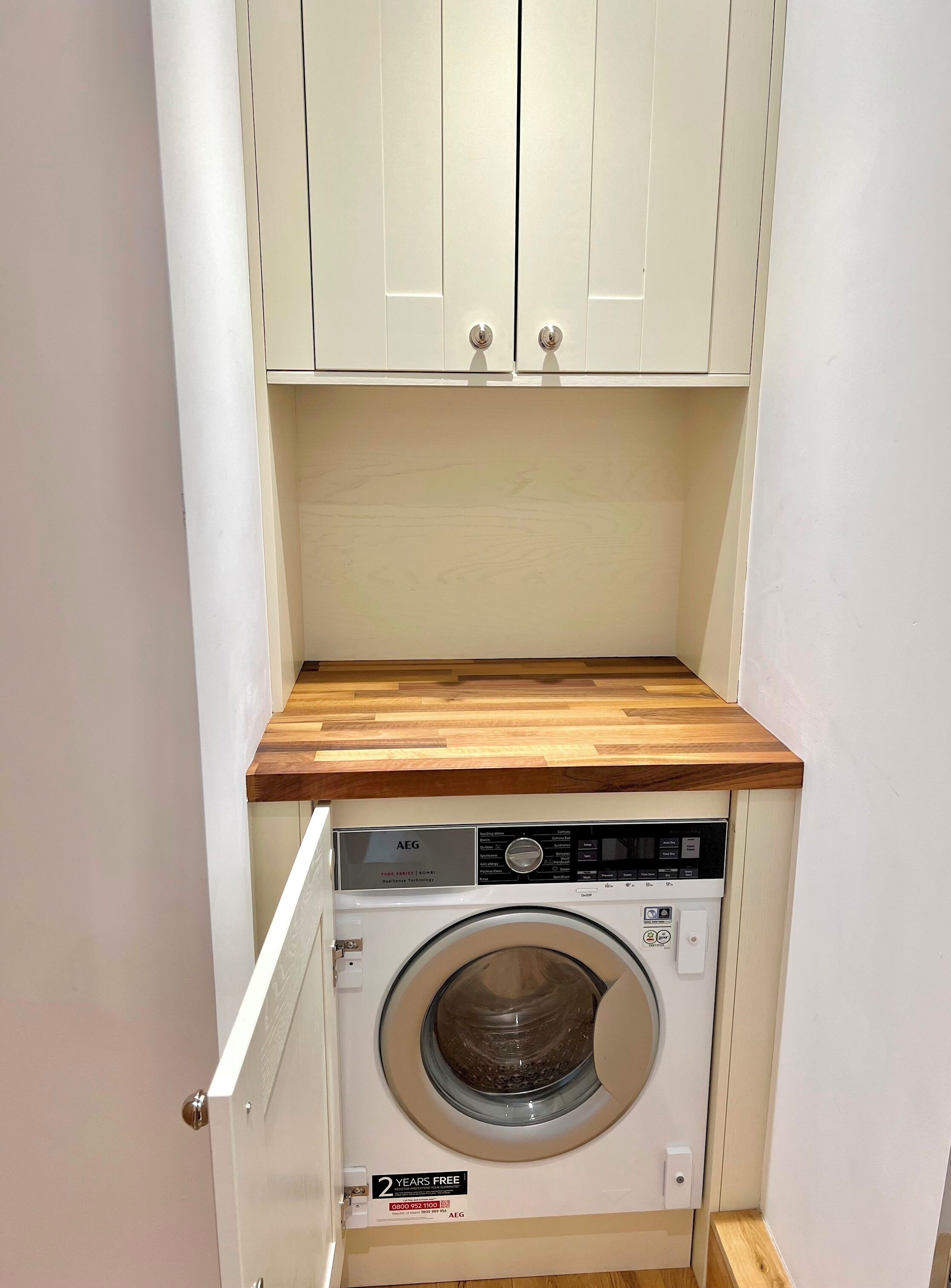 A compact laundry room with a built-in washing machine, wooden countertop, and white cabinets.