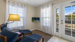 A room with a wooden floor, a sofa, a lamp, a painting, and a door leading to an outdoor area with a view of palm trees. - Daisy House: Downtown Cedar Key.  Walk to everything! (Cedar Key)