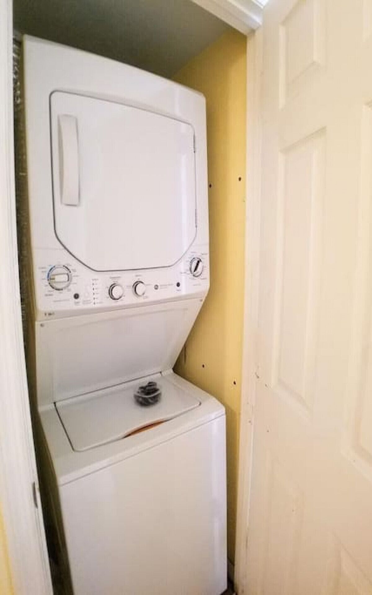 Un appareil compact combinant lave-linge et sèche-linge, installé dans une petite buanderie avec un mur jaune et une porte blanche.