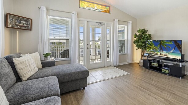 A living room with a grey sofa, a television on a black stand, and a large window with white curtains.
