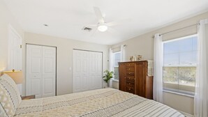 A bedroom with a bed, a ceiling fan, two white doors, a wooden dresser, and a window with blinds.