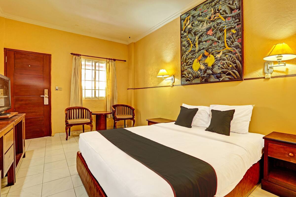 A hotel room with a large bed, two bedside tables, a desk with a chair, a television, and a painting on the wall.