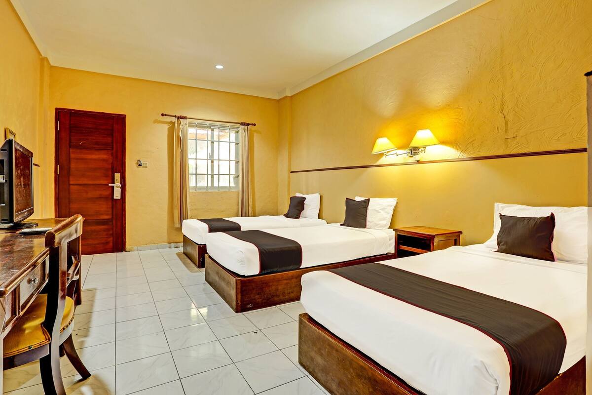 A hotel room with two beds, a desk with a television, a window with curtains, and a wall-mounted lamp.