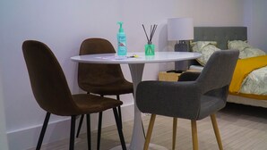 Ein Hotelzimmer mit einem runden Tisch, zwei Stühlen, einem Bett mit einer gelb-weißen Decke und einer Lampe, sowie einer Handdesinfektionsflasche.