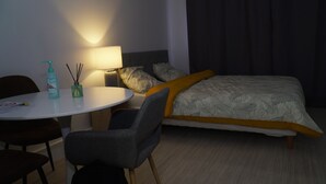 Ein Hotelzimmer mit einem Bett, einem runden Tisch und einem Stuhl. Auf dem Tisch steht eine Lampe und eine Flasche Handdesinfektionsmittel.