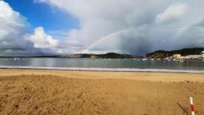 Une plage avec un arc-en-ciel, un rivage sablonneux et des bateaux sur l’eau.