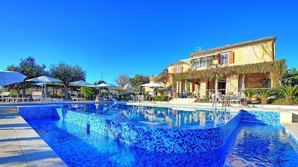 Ein Swimmingpool mit Mosaikfliesen, umgeben von Liegestühlen und Sonnenschirmen, und ein Gebäude mit einem bepflanzten, gefliesten Dach.