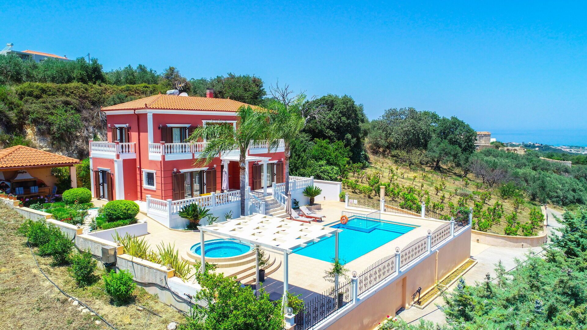 A two-story villa with a swimming pool, surrounded by lush greenery and a view of the sea.