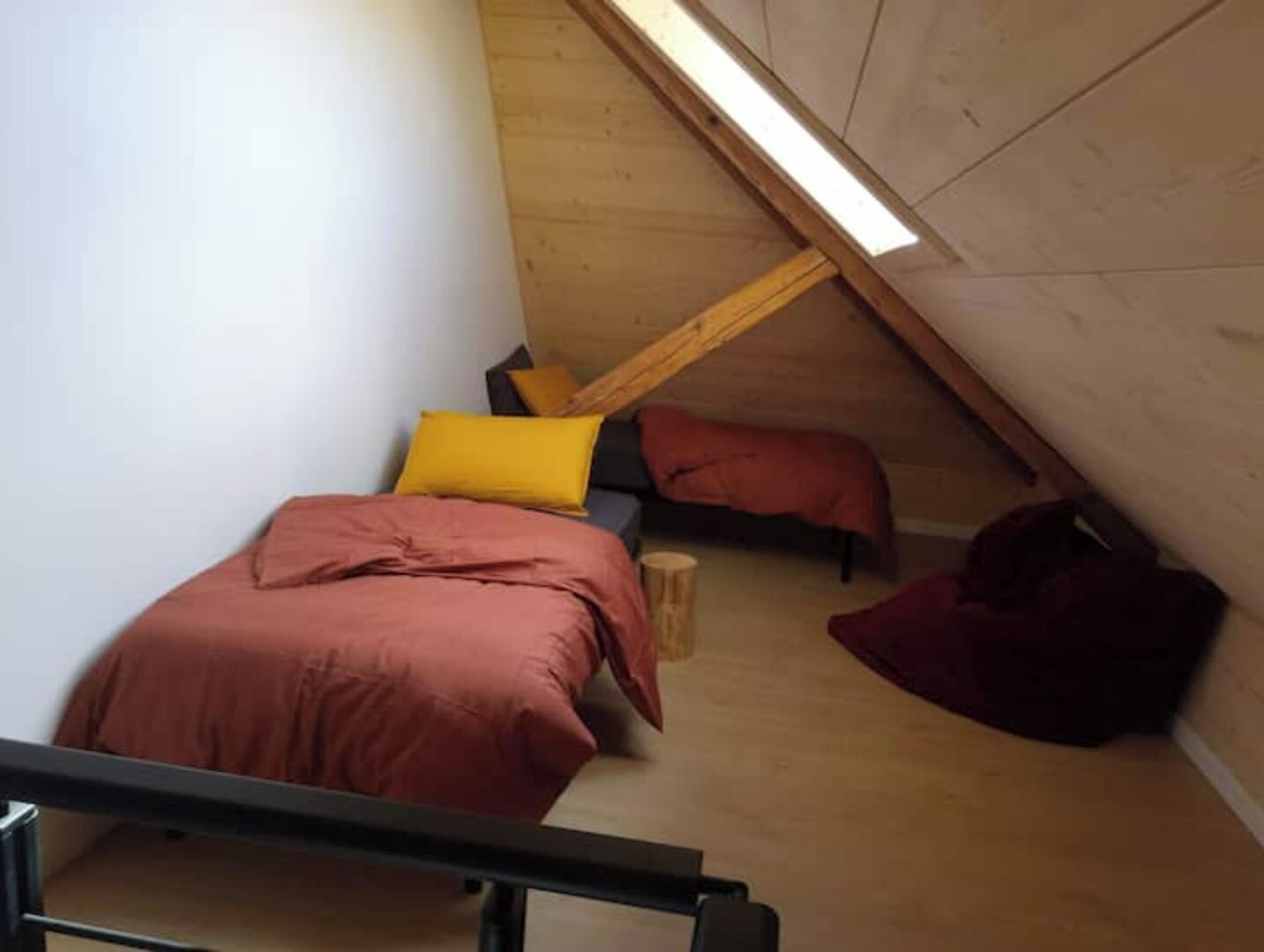 Une petite chambre mansardée, chaleureuse, avec un plafond en pente, un lit simple avec une literie rouge, un tabouret en bois et une chaise rouge.