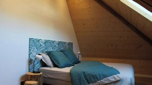A bedroom with a sloped ceiling, a bed with a blue bedspread, and a wall-mounted headboard with a leaf-patterned artwork. - Grand Appartement Avec Loggia (Orchamps-Vennes)