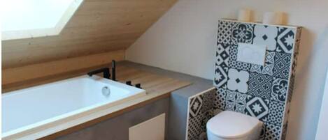 A small, modern bathroom with a sloped ceiling, a bathtub, a toilet, and patterned tiles.